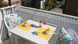 Un balcon avec une table blanche dressée pour deux personnes, agrémentée de sets de table jaunes et d’objets décoratifs tels qu’une étoile de mer et un coussin à l’effigie d’un flamant rose.