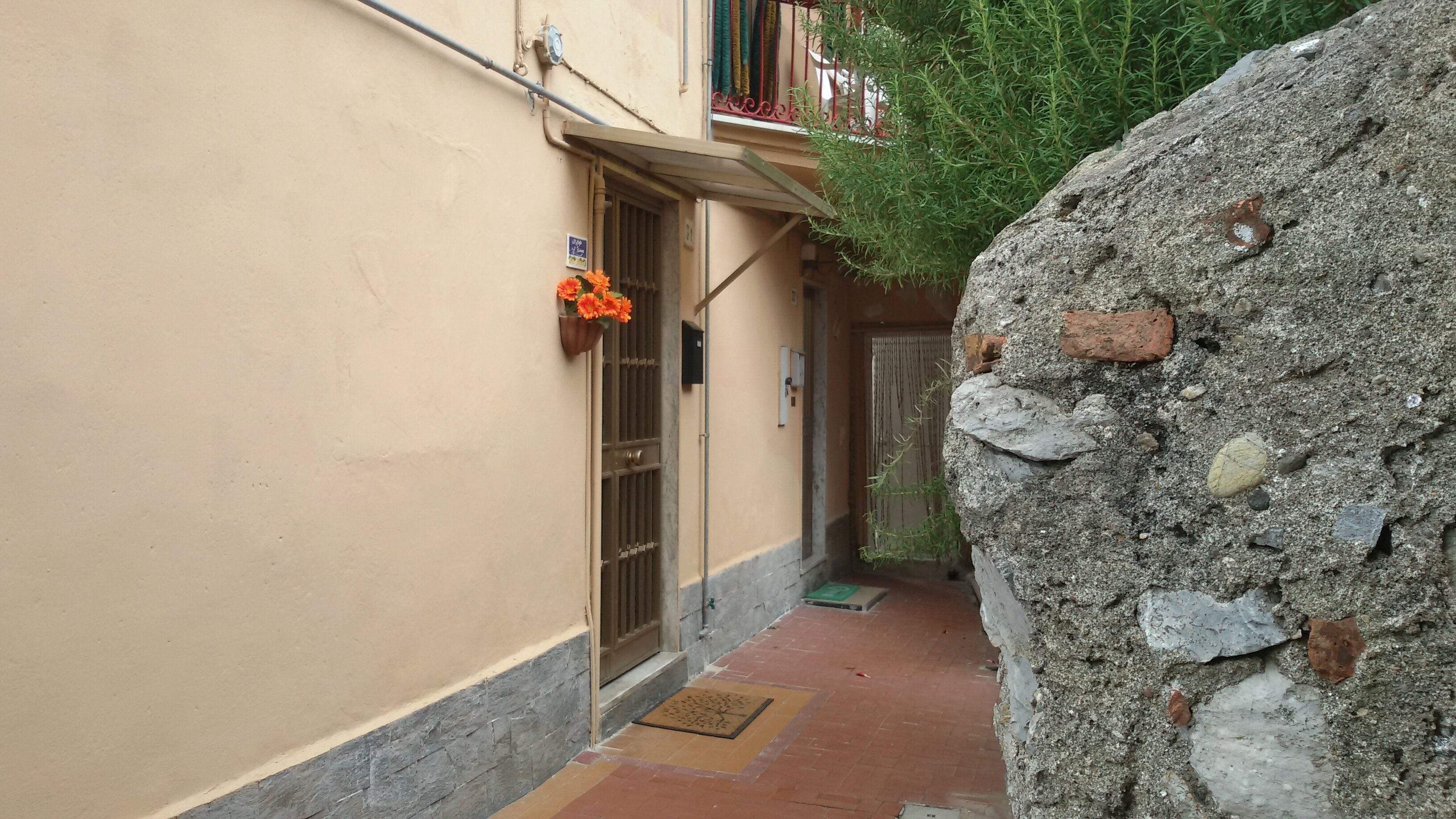 Un stretto vicolo con un muro in pietra su un lato e un edificio beige sull'altro. Si vede una piccola porta con un cancello in metallo, e un cesto di fiori è appeso alla parete.