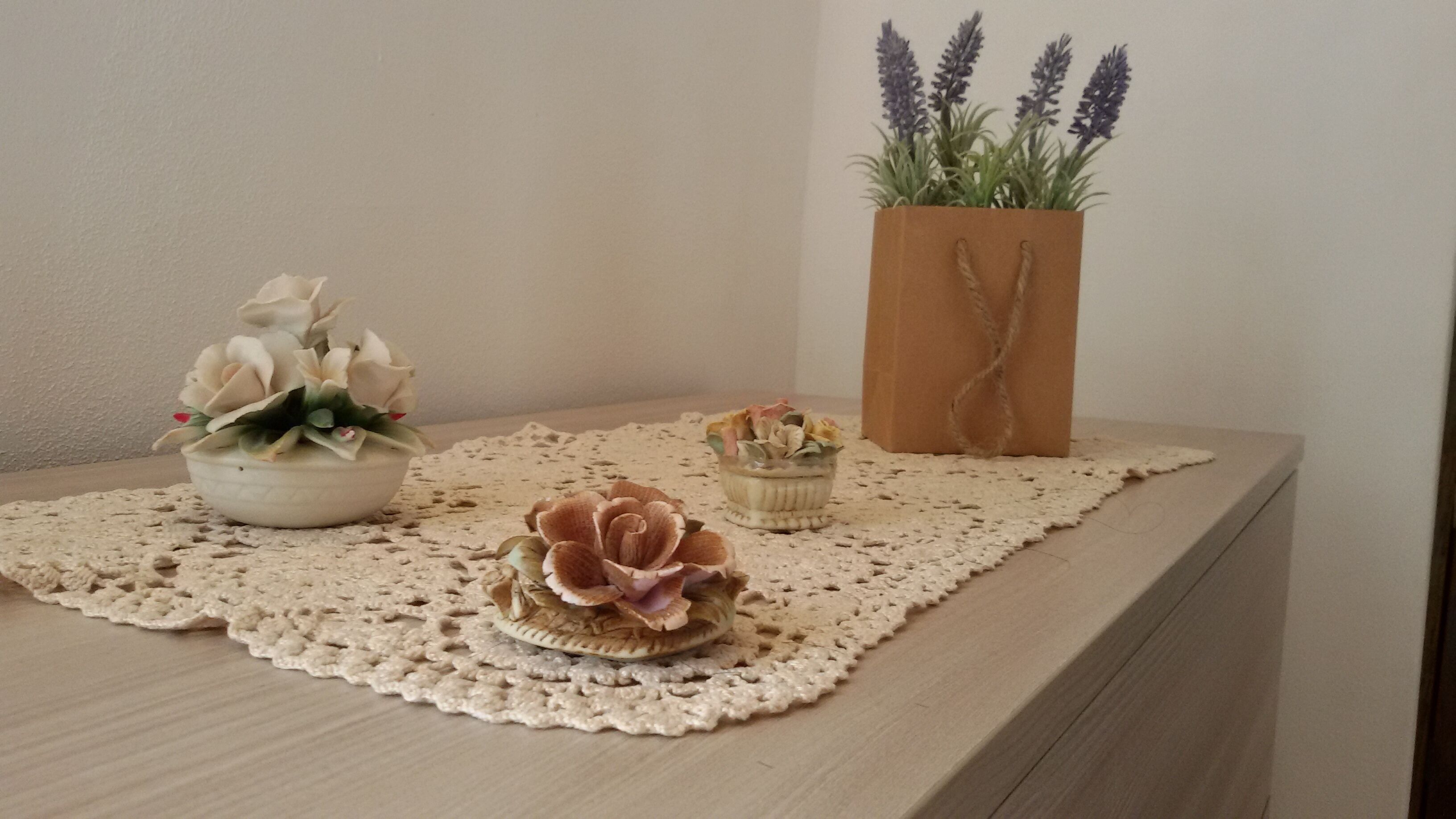 A wooden table with a doily, a white pot with flowers, a small bowl with flowers, and a brown paper bag with plants.
