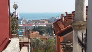 Un escalier étroit en pierre mène à un niveau supérieur, et il est entouré de bâtiments traditionnels aux toits de tuiles rouges, d’où l’on peut apercevoir la mer au loin.