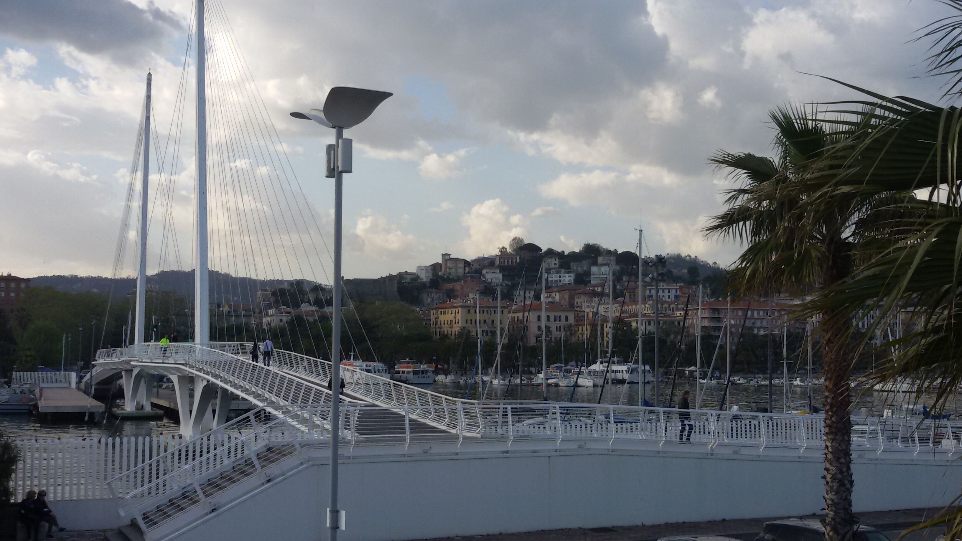 Eine Fußgängerbrücke mit weißer Geländer, eine Straßenlaterne und Blick auf einen Hafen mit Booten und Gebäuden im Hintergrund.