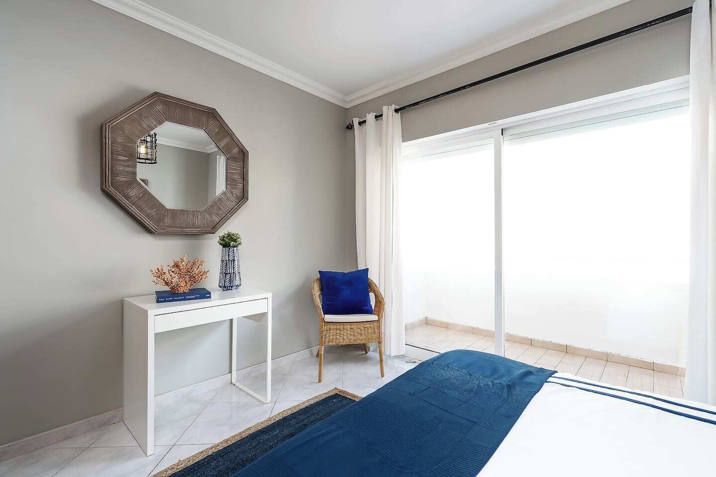 A modern bedroom with a large window, a white console table, a wicker chair, and a blue bedspread.