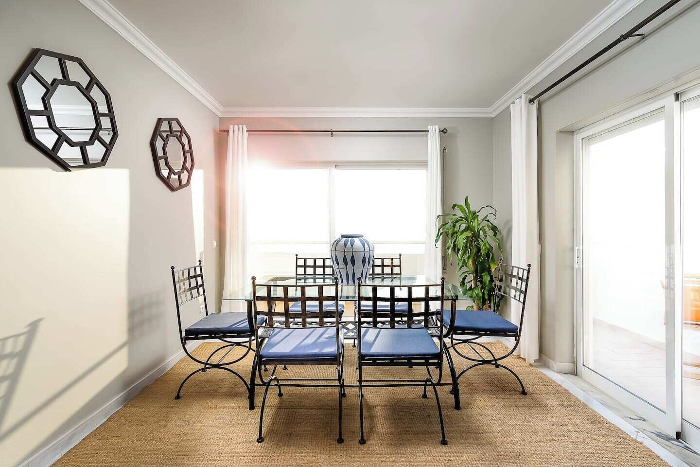 Un comedor con mesa de vidrio y sillas de metal, un ventanal con cortinas y una planta en maceta.