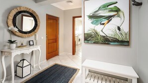 A hallway with a white console table, a mirror with a woven frame, a framed bird painting, and a wooden door. - Comfortable 2-bed apartment, sleeps 6. Tastefully decorated and spacious. Perfect base to explore the Algarve — Lagos, Albufeira and beyond. Free street parking. Easy self check-in. Always on hand to help. (Portimão)