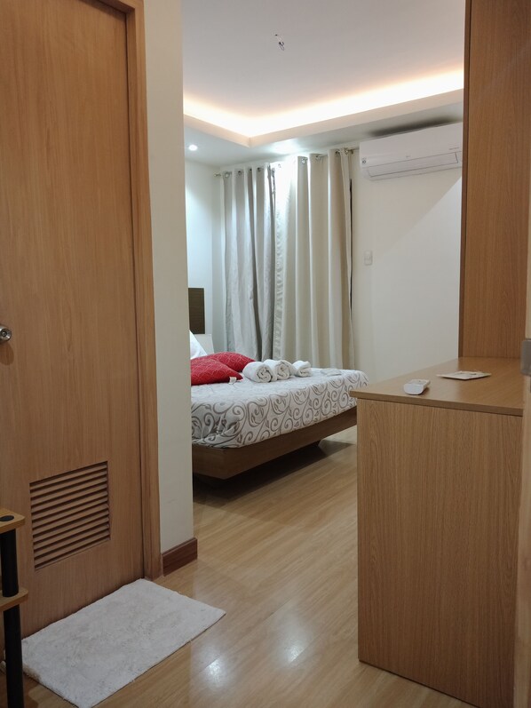 A hotel room with a wooden desk, a bed with patterned bedding, a white rug, and a ceiling-mounted air conditioner. - FGR Vacation Home at Mivesa Garden Residences (Cebu City)