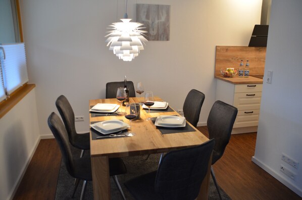 Un coin repas avec une table en bois dressée pour quatre personnes, entourée de chaises, d’un luminaire suspendu moderne et d’un petit espace cuisine comprenant un comptoir et des placards.