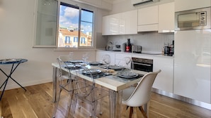 A modern kitchen with a dining area, featuring a large window, white cabinetry, and a stainless steel refrigerator.