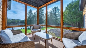 Une pièce chaleureuse, meublée de mobilier en osier, dotée de grandes fenêtres et offrant une vue sur un lac entouré d’arbres.