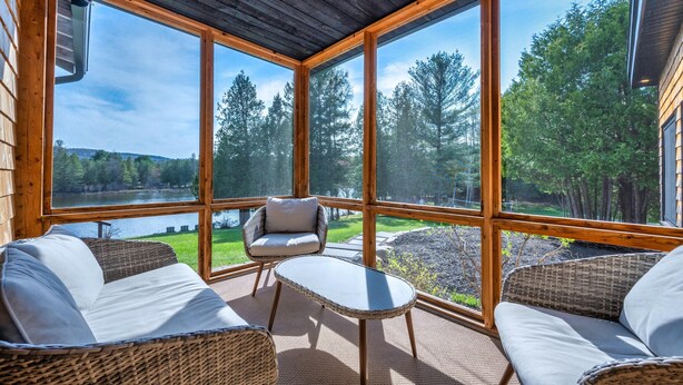 Ein gemütliches Zimmer mit Korbmöbeln, großen Fenstern und Blick auf einen von Bäumen umgebenen See.
