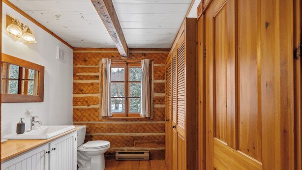 Une salle de bain rustique, avec des murs et un plancher en bois, un lavabo blanc, des toilettes et une fenêtre agrémentée de rideaux.
