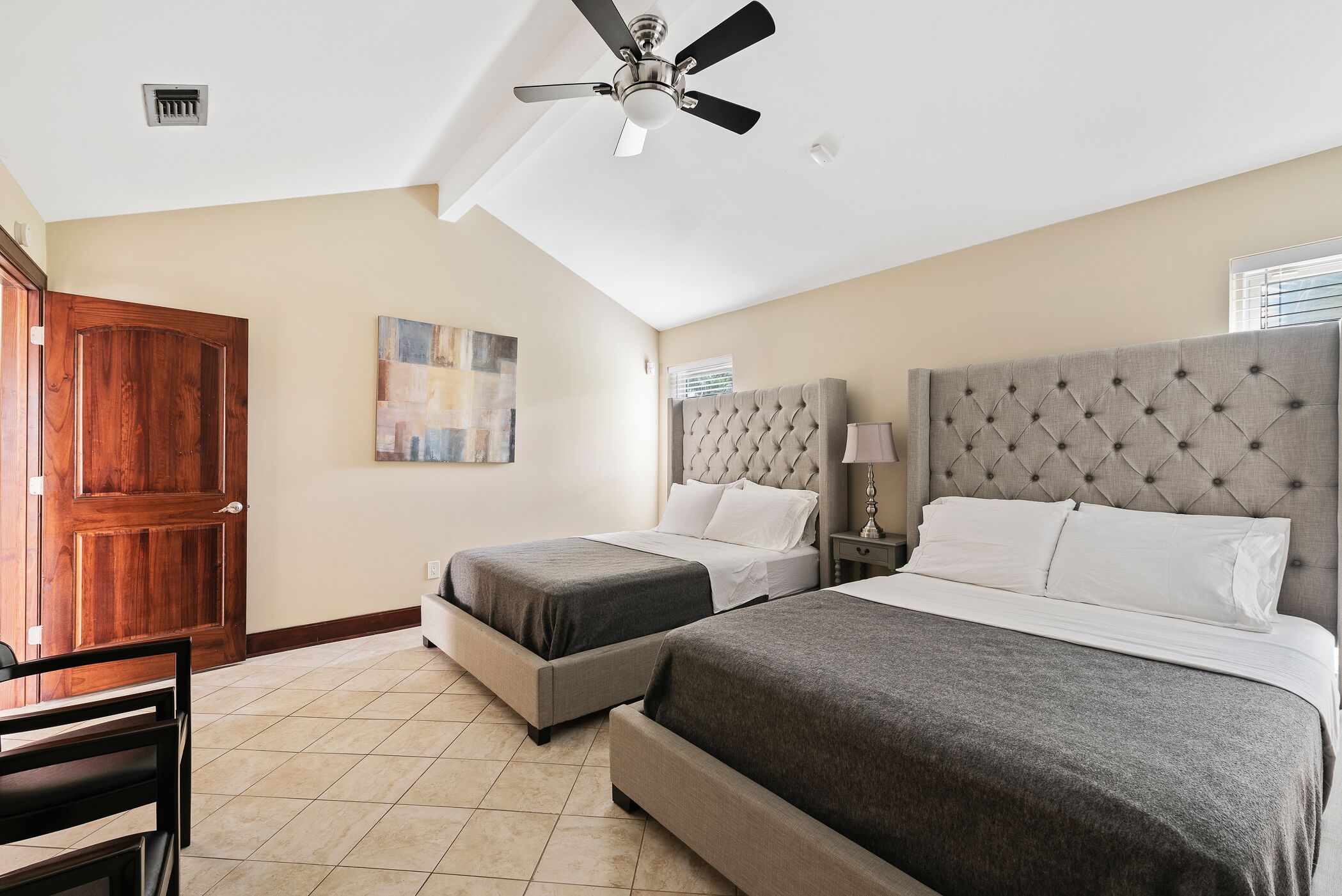 A hotel room with two beds, a ceiling fan, a wooden door, and a painting on the wall.