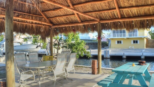 A covered outdoor seating area with tables and chairs, a thatched roof, and a view of a water body and buildings in the background. - Island Life at its best (Key Largo)