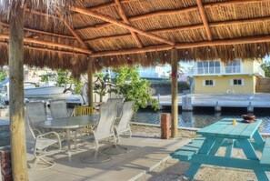 A covered outdoor seating area with tables and chairs, a thatched roof, and a view of a water body and buildings in the background. - Island Life at its best (Key Largo)