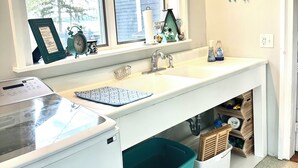Une cuisine avec une fenêtre, un évier et un plan de travail sur lequel se trouvent une bouilloire, une tasse et une petite plante. Une poubelle de recyclage se trouve sous le plan de travail.