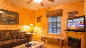 A cozy log cabin interior with a wooden sofa, a dining table, a fireplace, and a television displaying a news broadcast. - Private, Super Fast WiFi Free, NFL Sunday, VideoArcade, Fishing & 2 Pools, Gated (Sevierville)