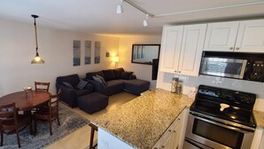 A compact kitchen with a microwave, oven, and granite countertop. There is a dining area with a wooden table and chairs, and a dark-colored sofa in the living area. - Cape May Condo Getaway w/ Rooftop Deck, Onsite Parking, 1/2 Block to the Beach (Cape May)