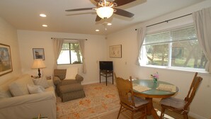 A living room with a sofa, armchair, and dining table with chairs. There is a ceiling fan, a TV, and a window with curtains.