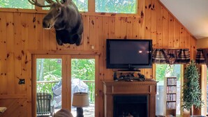 Un acogedor interior de cabaña con un amplio techo de madera, una cabeza de alce montada, una chimenea, un televisor y una estantería.