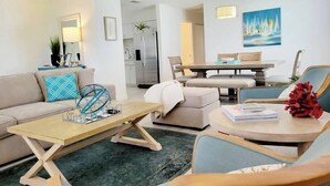 Ein modernes Wohnzimmer mit einer Couch, einem Couchtisch und Sesseln, ein Essbereich mit Tisch und Stühlen sowie eine Küche mit Kühlschrank.
