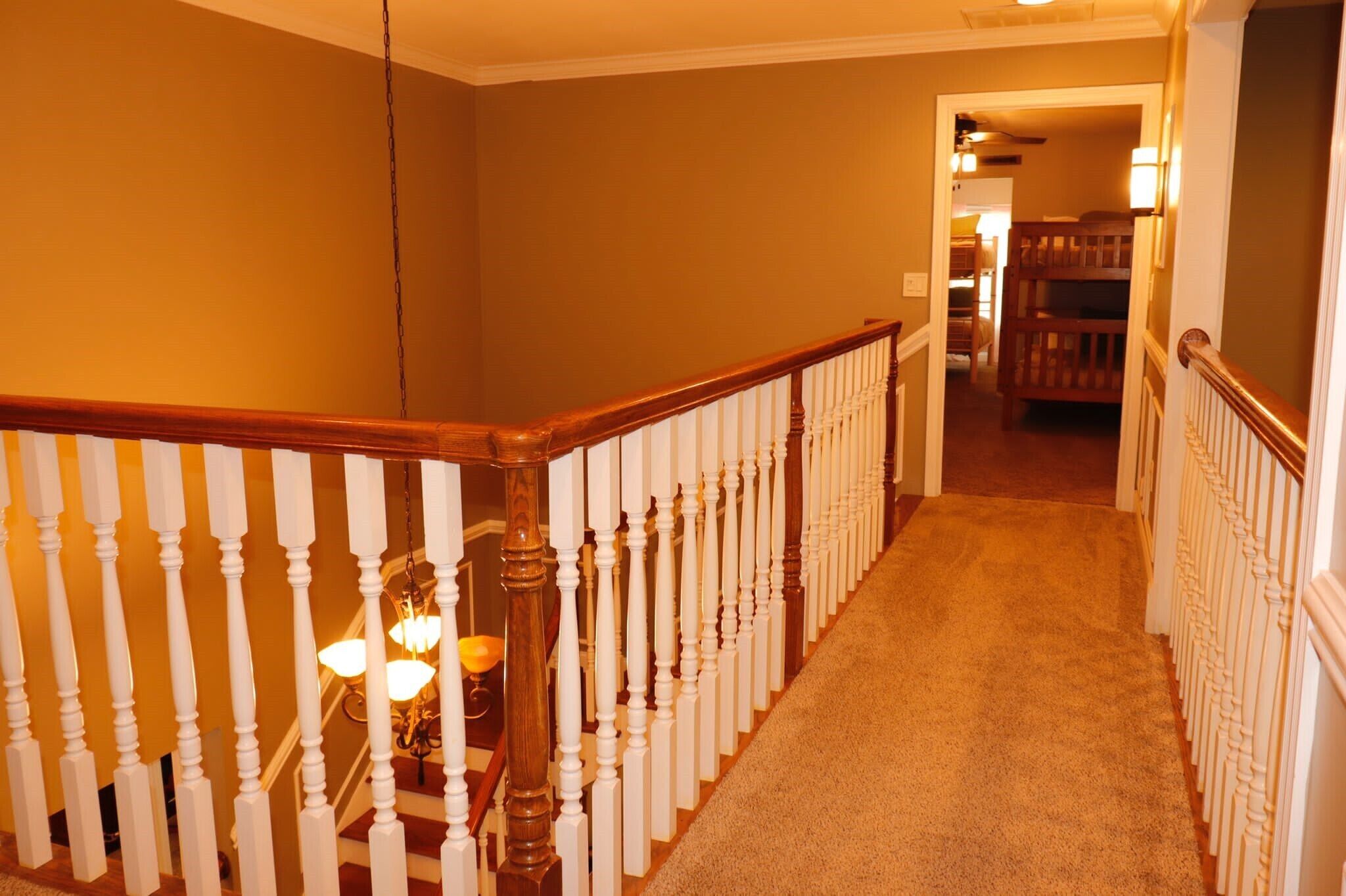 A carpeted staircase with white balusters and a wooden handrail, leading to a room with a bunk bed and a lamp.