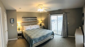 A bedroom with a bed, nightstands, a ceiling fan, a painting, and a view of houses through a window.