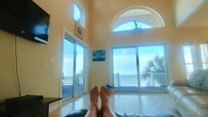 A living room with a large window overlooking the ocean, a flat-screen TV mounted on the wall, and a sofa with cushions. - Beachfront, kid friendly seas, seclusion, nature (Carrabelle)