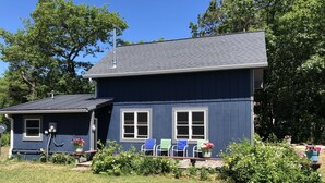 Une maison à deux étages, à l’extérieur d’un bleu foncé, avec un petit garage attenant et un porche à l’avant, agrémenté d’un banc et de plantes en pot.