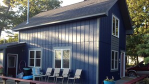 Une maison bleue à deux étages, dotée d’un porche et d’un espace de repos couvert avec des chaises, et un véhicule garé à proximité.