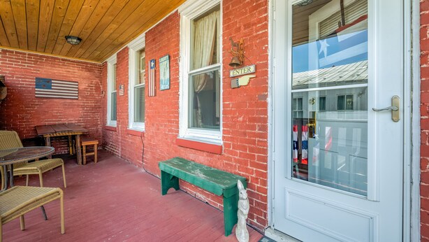 A covered porch with a wooden ceiling, red brick walls, and a white door. There are wicker chairs, a wooden bench, and a small table. -  Lincoln Quarters, 2nd floor,  Only 2 blocks to the center of town. (Gettysburg)