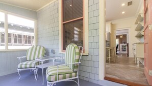 A covered patio area with two striped chairs and a table, a large window with blinds, and a view into the house. - HISTORIC DISTRICT ~ Free Parking! (Saint Augustine)