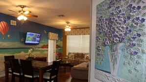 A dining area with a wooden table and chairs, a television mounted on the wall, and a ceiling fan.