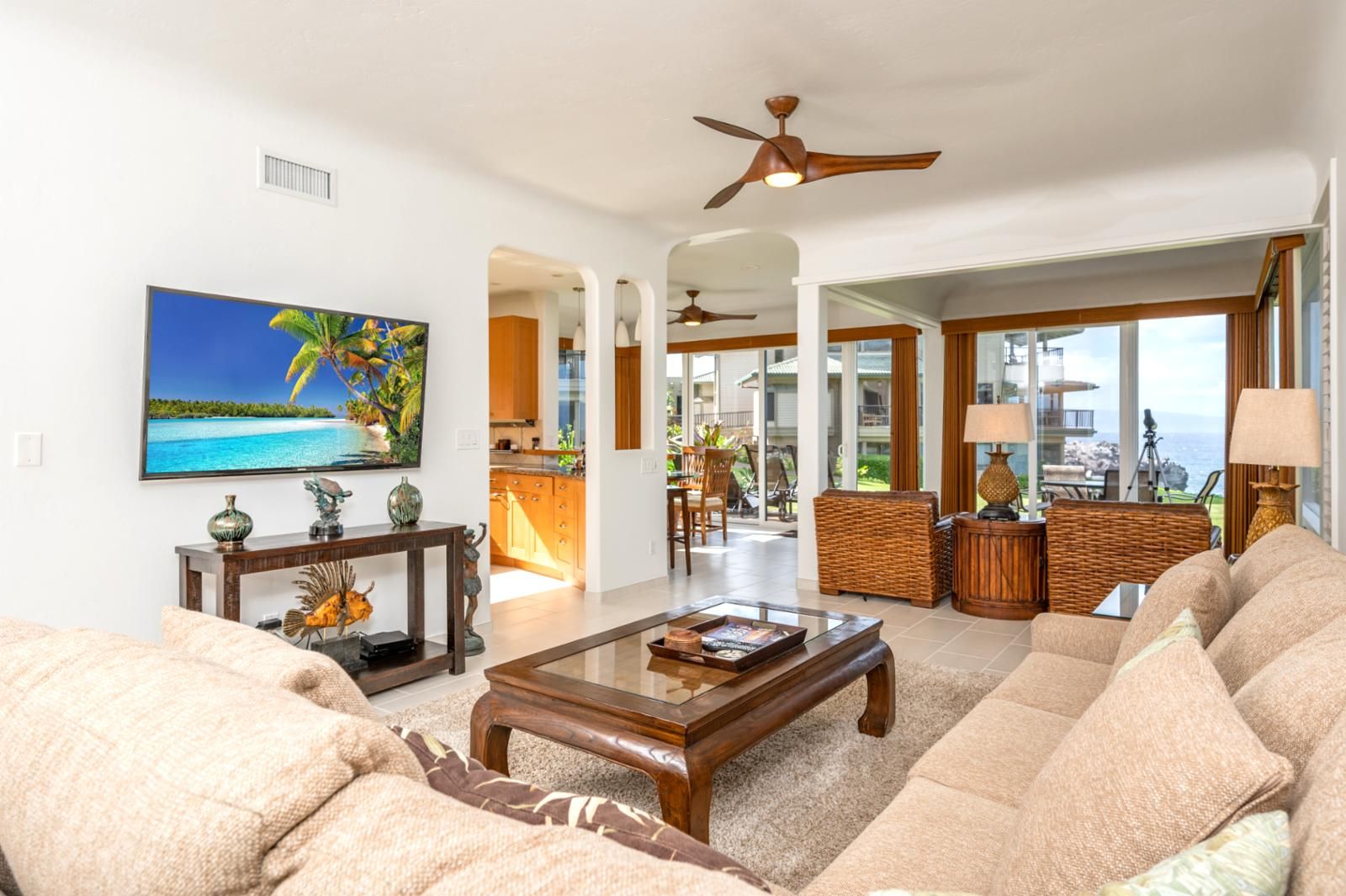 A spacious living room with a large flat-screen TV, a wooden coffee table, and a comfortable beige sofa. There is a ceiling fan, a dining area with a view of the ocean, and a kitchen visible through an open doorway.