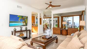 A spacious living room with a large flat-screen TV, a wooden coffee table, and a comfortable beige sofa. There is a ceiling fan, a dining area with a view of the ocean, and a kitchen visible through an open doorway.