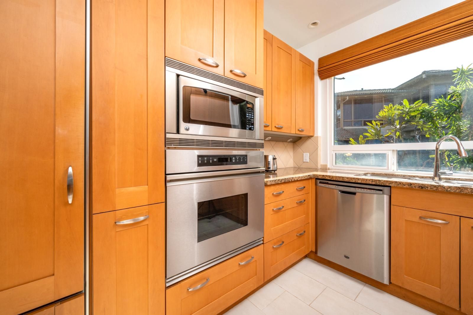 A modern kitchen with wooden cabinets, stainless steel appliances, a window with a view of greenery, and a built-in dishwasher.