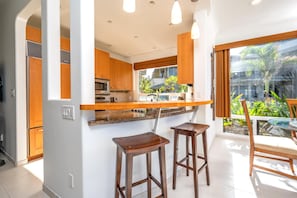 Une cuisine moderne avec des armoires en bois, un comptoir en granit et un four encastré. On y trouve deux tabourets en bois et une table en verre avec des chaises.