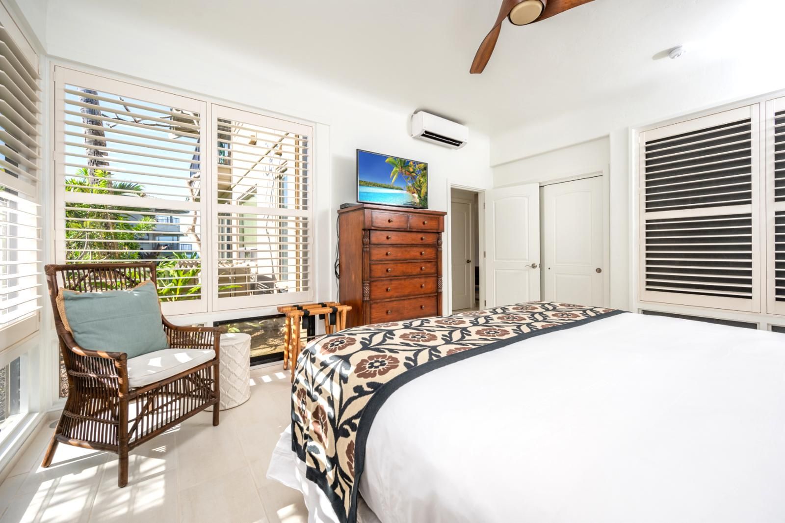 A bedroom with a large bed, a wooden dresser, a wicker chair, and a view of the outdoors through the window.