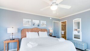A bedroom with a large bed, a ceiling fan, a nightstand with a lamp, a bench, and a mirror. - BEACH ACCESS! Lost Colony Villas Beach home with multiple relaxing outdoor deck (Port Aransas)