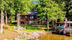 A cabin with a stone foundation, wooden deck, and a dock extending into a river. - Waterfront Cozy Log Cabin Lake House - The Sunset Cabin (Leesville)