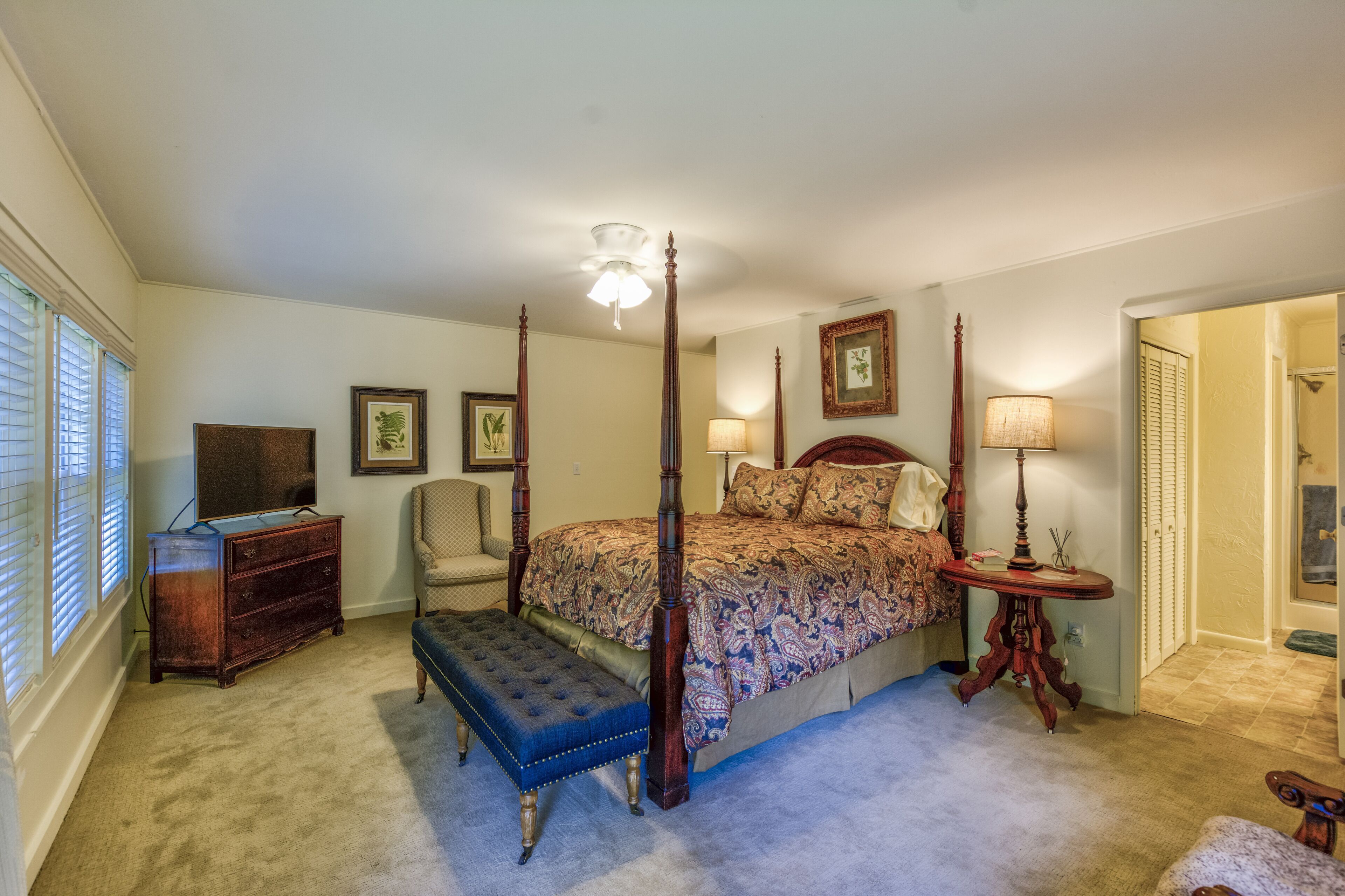 A four-poster bed with a canopy, a wooden dresser, a bench, and a small table with a lamp.