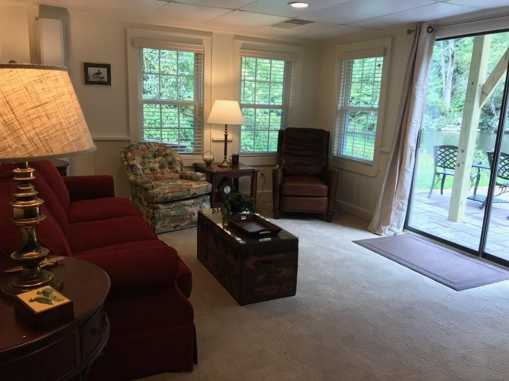 Un salon avec un canapé rouge, un fauteuil à motifs floraux, une table basse en bois et une lampe. Il y a deux fenêtres équipées de stores, une porte donnant sur un patio et une vue sur la verdure à l’extérieur.