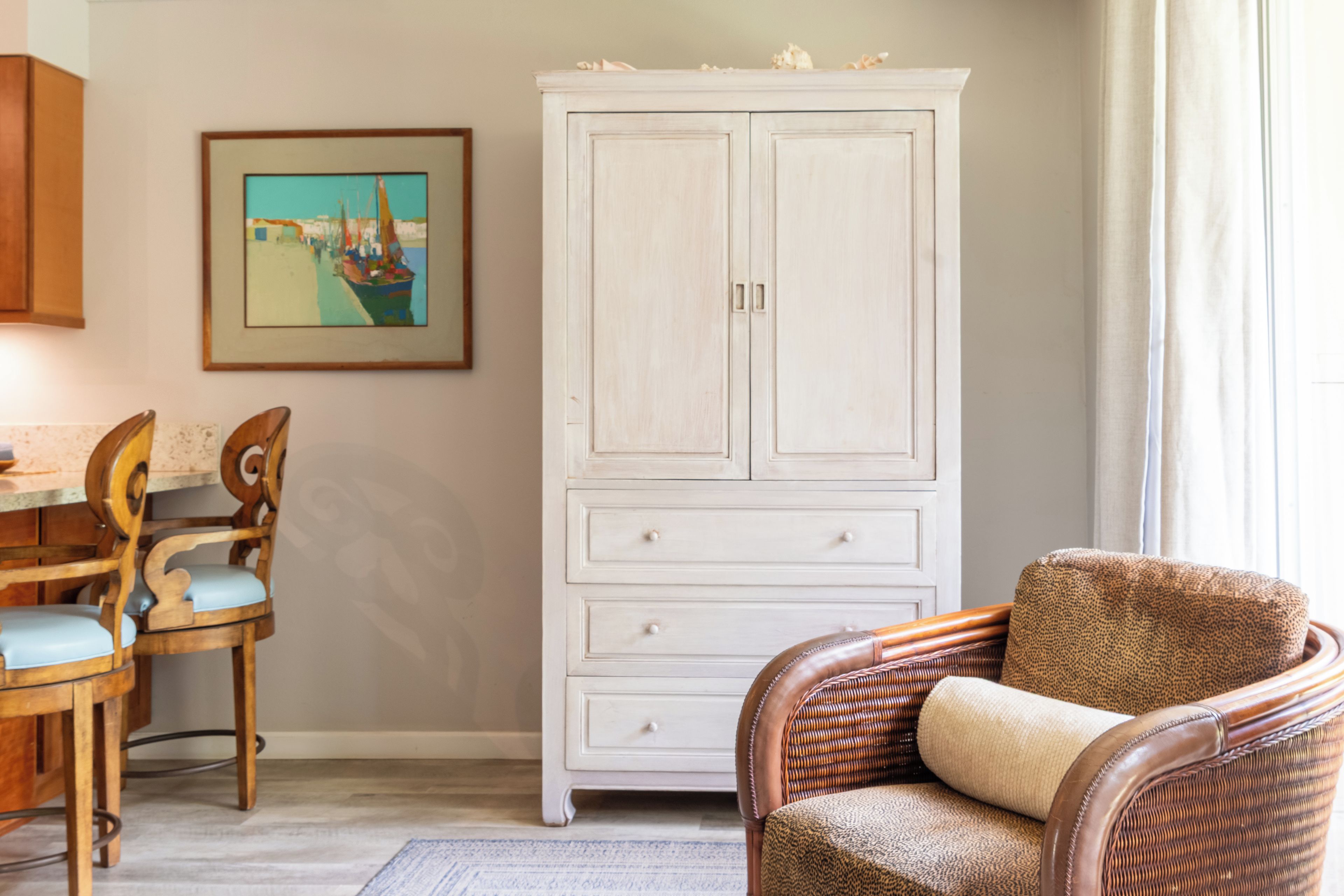 Ein Zimmer mit einem weißen Schrank, einem Korbstuhl, einem Holzesstisch mit Stühlen und einem gerahmten Gemälde an der Wand.