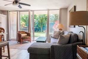 Una habitación con ventilador de techo, una silla de mimbre, un sofá, una mesa y vistas a un jardín a través de puertas de vidrio.