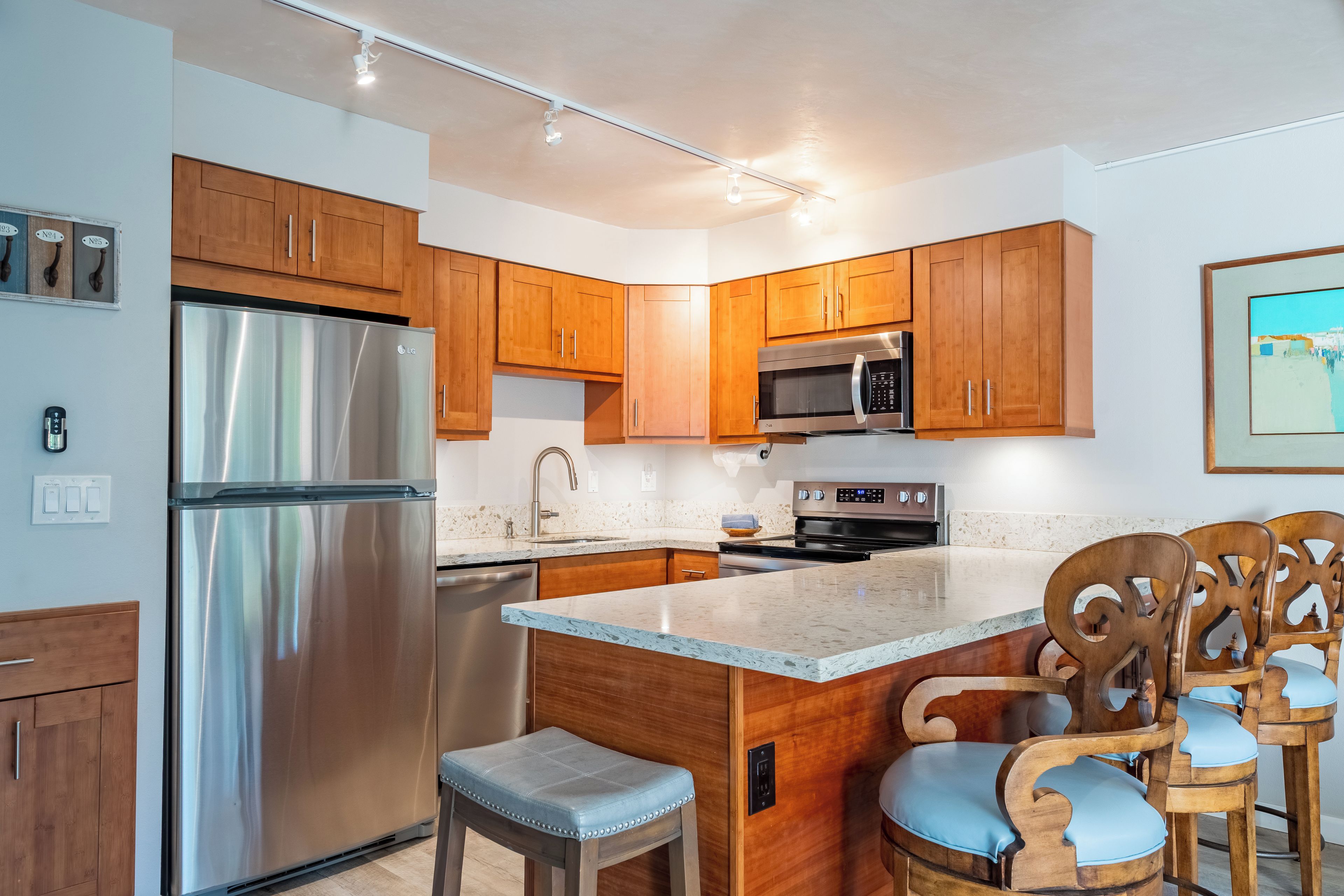 A modern kitchen with wooden cabinets, stainless steel appliances, a marble countertop, and a seating area with wooden chairs.