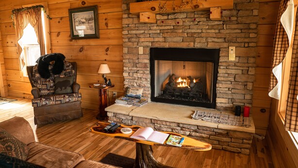A cozy cabin interior with a stone fireplace, wooden walls, a cushioned armchair, a coffee table with books and a remote control, and a plaid-patterned throw on the armchair. - Honeymoon/couples Retreat -  Private Custom Built Log Cabin - Wi-fi (Sevierville)