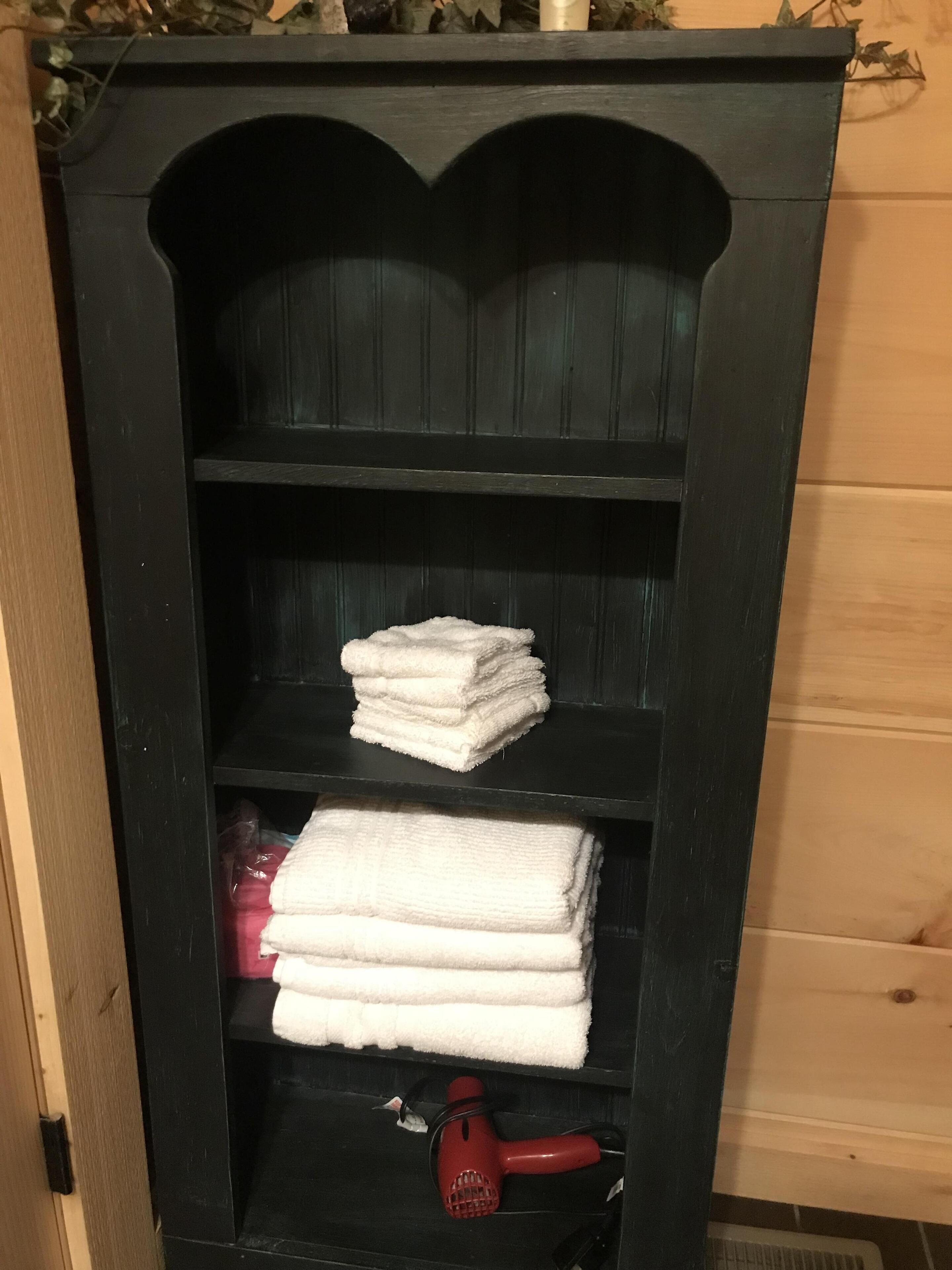 A black wooden cabinet with shelves holding folded white towels and a red hairdryer.