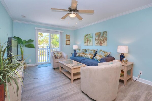 A living room with a blue wall, a ceiling fan, a sofa, a coffee table, and a side table with a lamp. - Oceanview Condo OW 6103 in Oak Island, NC – perfect for 4 guests! (Oak Island)