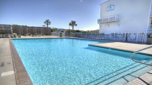 Une piscine aux eaux cristallines entourée d’une terrasse carrelée, un bâtiment blanc orné d’un logo et, au fond, une clôture en bois.