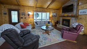 L’intérieur d’un chalet confortable, avec une cheminée, des murs en bois, un canapé à carreaux, des fauteuils en cuir et un téléviseur fixé au mur.
