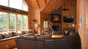 A cozy cabin interior with a stone fireplace, leather seating, a television, and large windows overlooking a forest. - SPACIOUS FAMILY RETREAT NEAR ALL THE FUN! STUNNING VIEW, EASY ACCESS. (Sevierville)