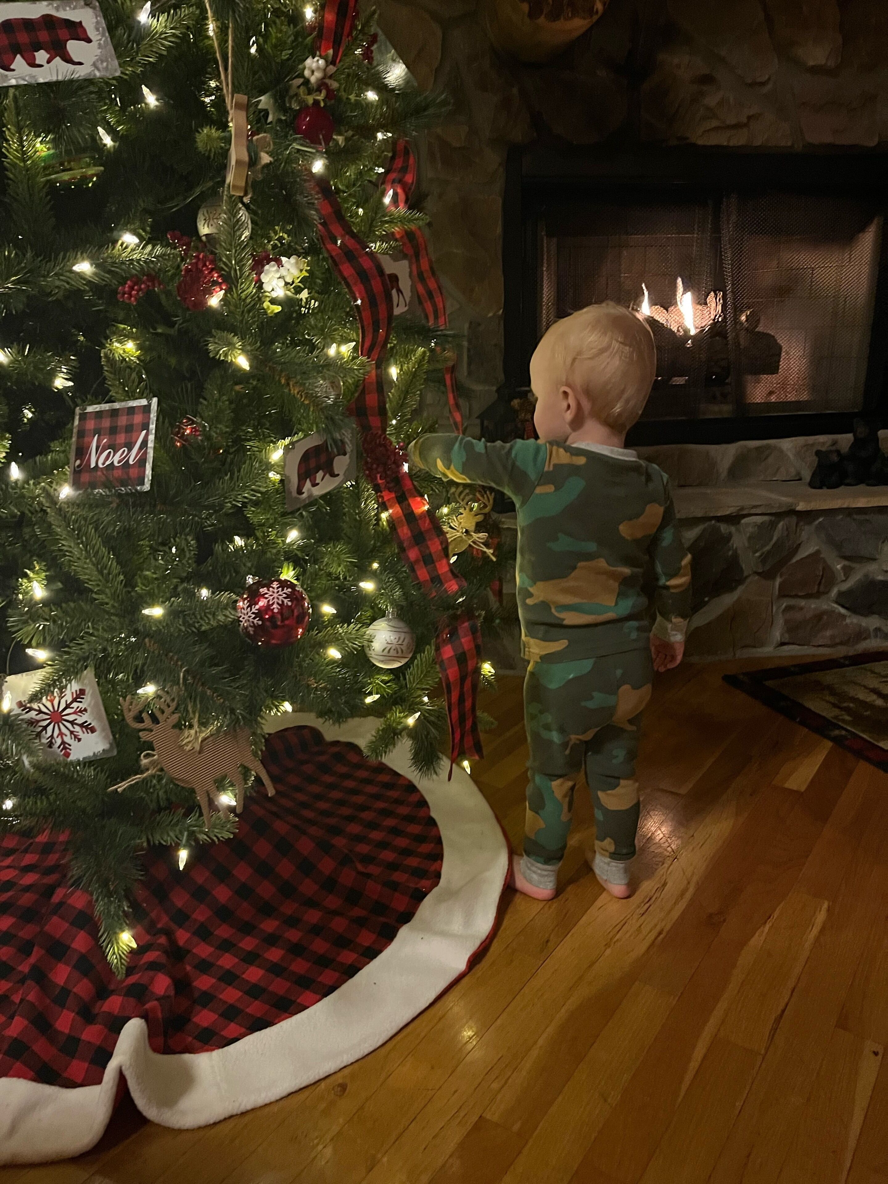 Un enfant se tient près d’un sapin de Noël décoré de rouge et de noir, avec une cheminée en arrière-plan et un couvre-pied à carreaux au pied de l’arbre.